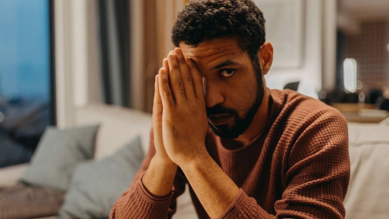 A man with a beard sits on a couch, covering his face with his hands.