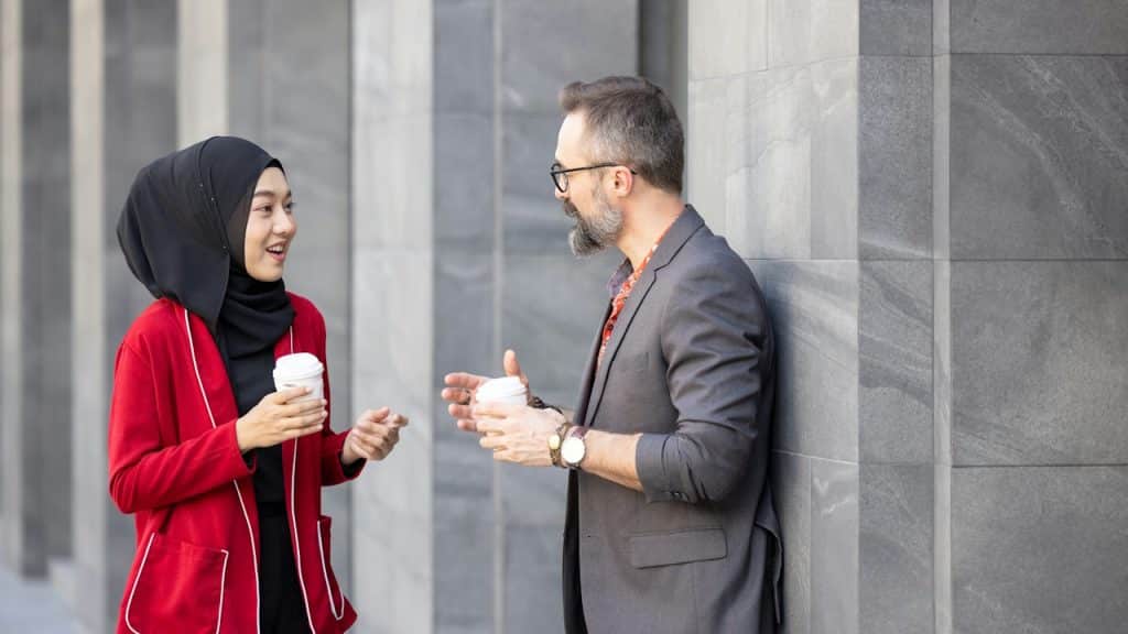 A woman wearing a hijab and a red blazer is standing and smiling while talking to a casually dressed man leaning against a stone column, both holding coffee cups.