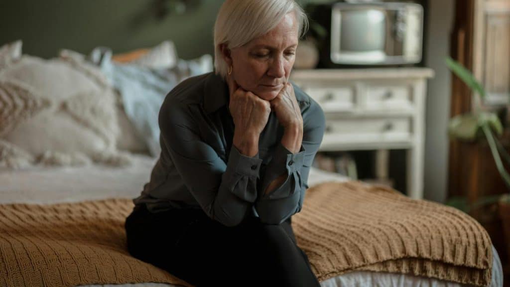 An elderly woman sitting on a bed with her hands under her chin, looking down thoughtfully.