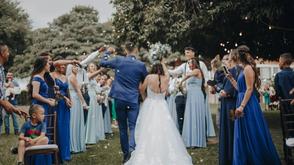 A bride and groom are walking away from the camera through a line of wedding guests who are cheering and throwing confetti or petals.