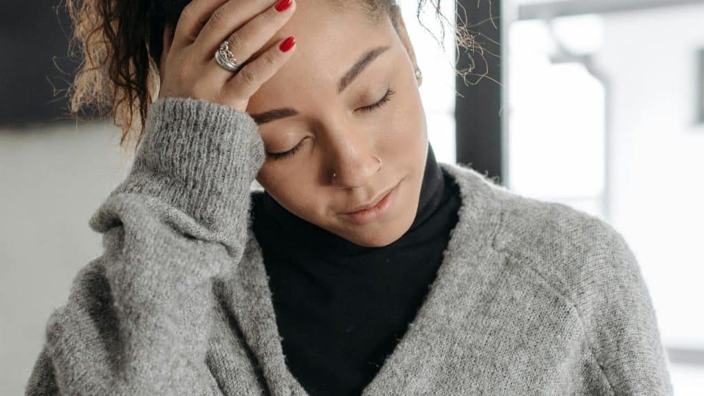 A woman in a gray sweater touching her forehead with her eyes closed.