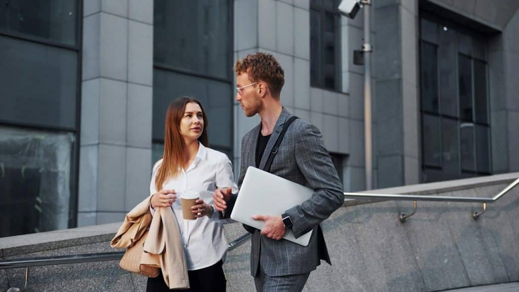 A woman holding a coffee cup walking with a man carrying a laptop outside an office building.