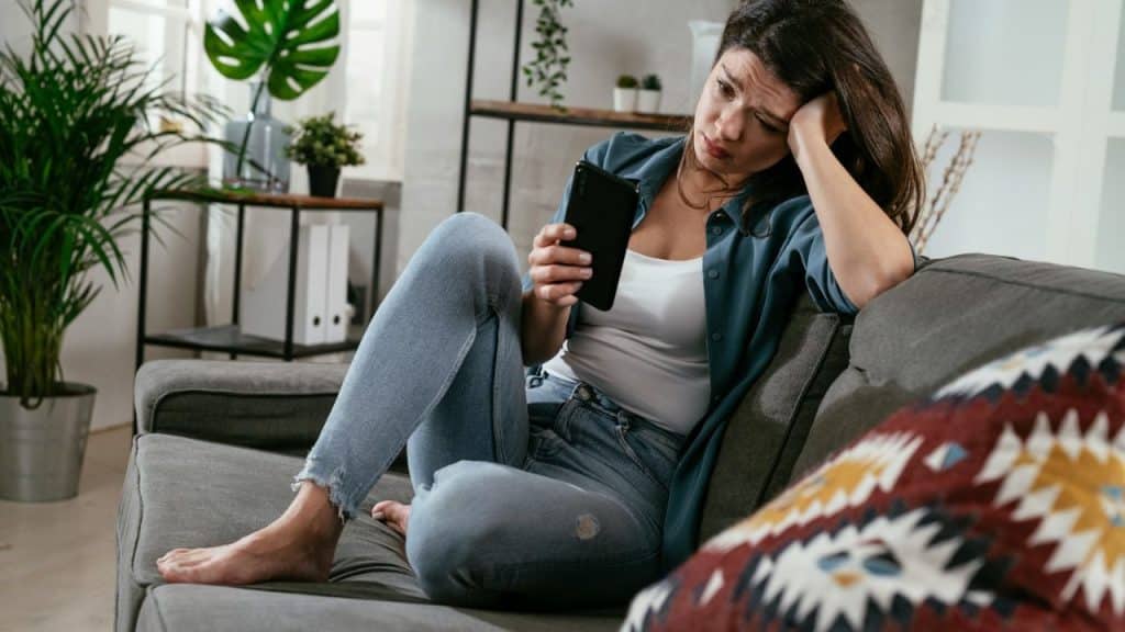 A distressed woman sitting on a sofa, looking at a phone while resting her head on one hand.