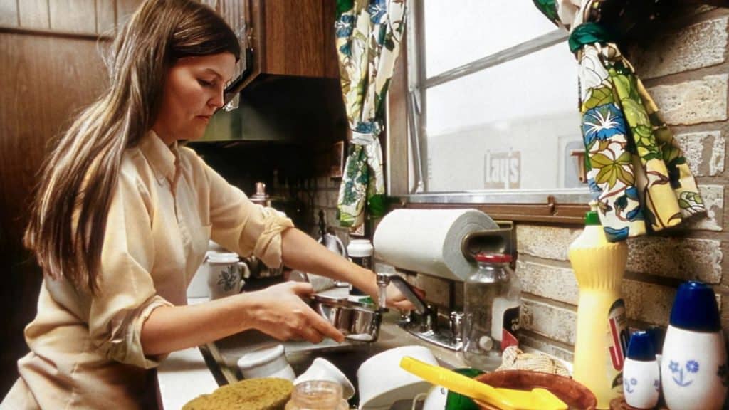 A woman with long brown hair washes dishes at a sink in a kitchen with brick walls.