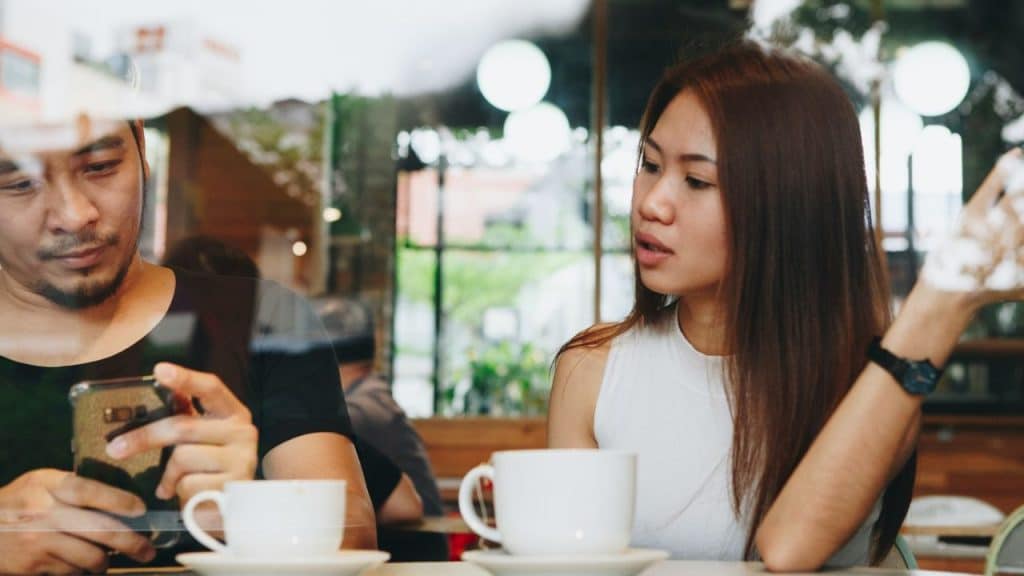 Man intently looks at his phone while a woman across from him talks in a coffee shop.