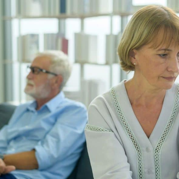 An older woman looking distant in the foreground; an older man sits in the blurred background.