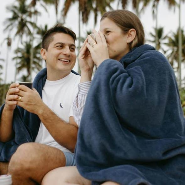 A couple wrapped in blankets sitting on a beach and drinking warm beverages.