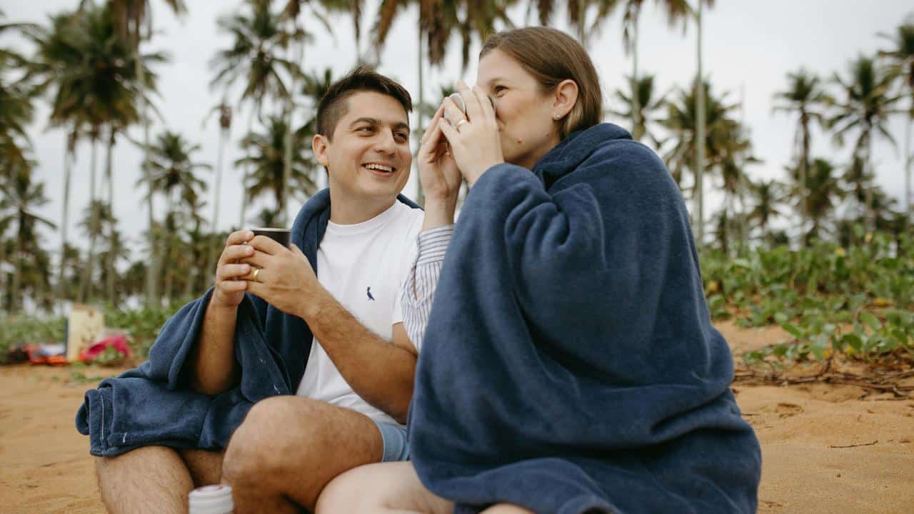 A couple wrapped in blankets sitting on a beach and drinking warm beverages.