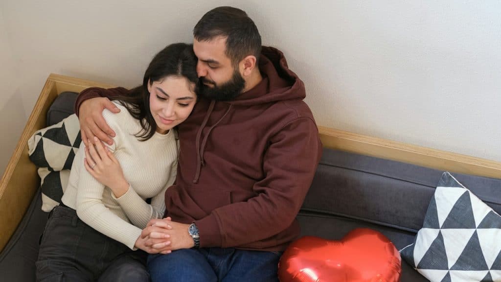 A couple is sitting closely together on a couch, embracing and holding hands, with a large red heart-shaped balloon next to them.