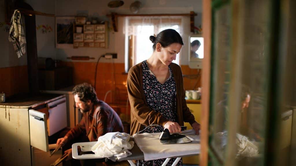 A woman is ironing clothes in a rustic, dimly lit kitchen, while a man is crouched down working near a stove in the background.