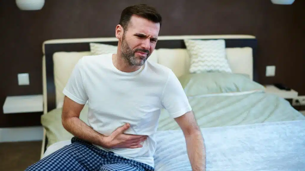 Man in a white shirt sitting on a bed clutching his stomach in pain.