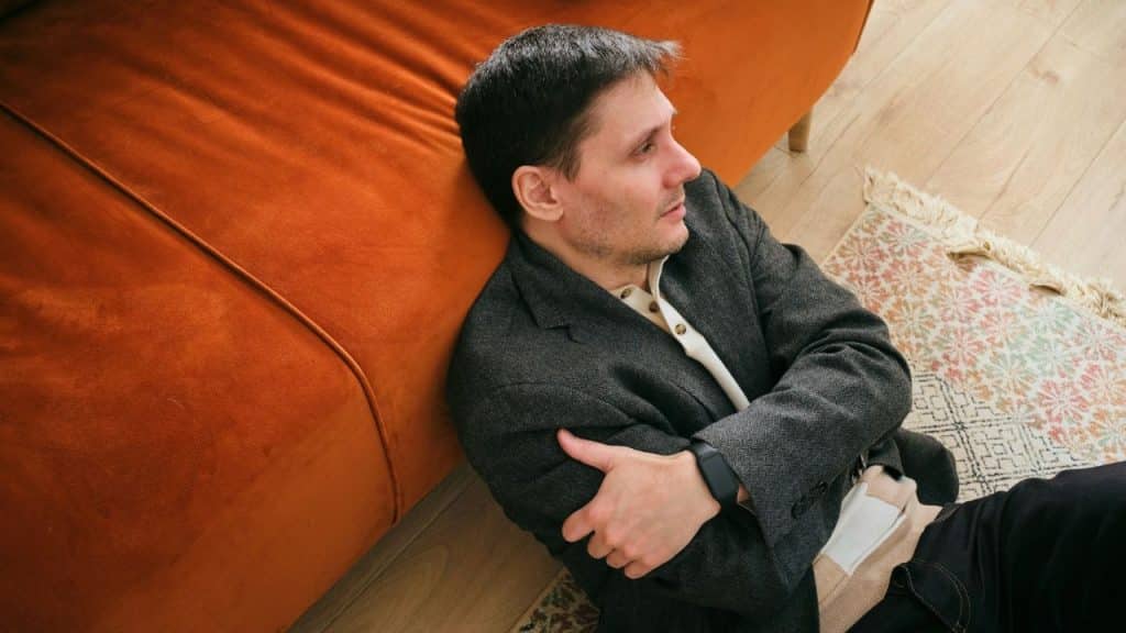 Man sitting on a wooden floor, leaning against an orange couch, hugging his knees.