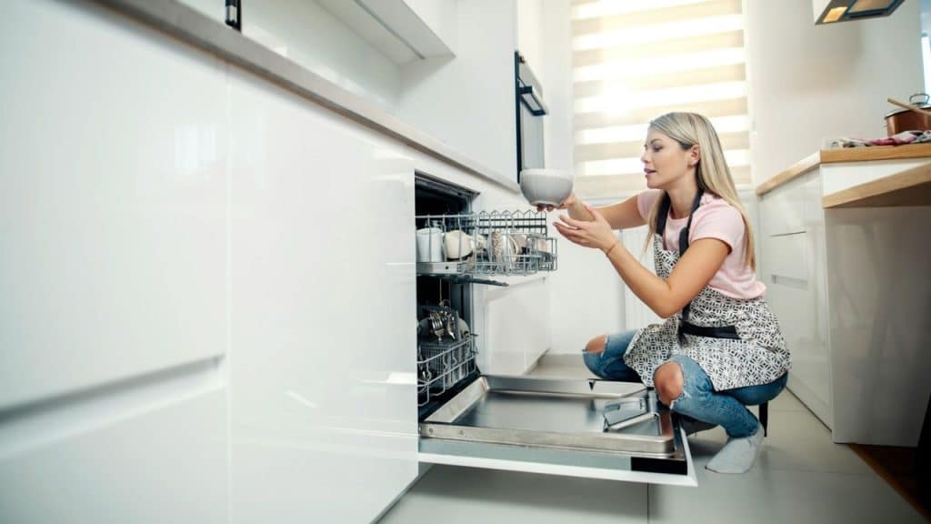 Blonde woman squatting, loading dishes into a modern white dishwasher.