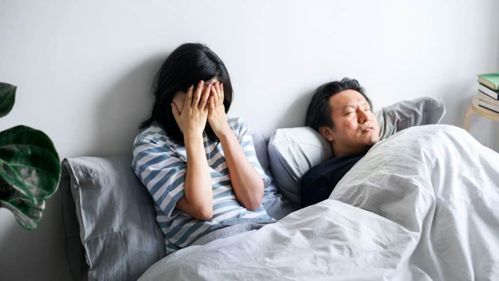 Woman crying with hands over her face next to a man sleeping in bed.