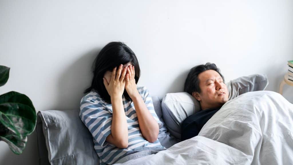 A distressed woman is sitting up in bed with her face covered by her hands, while a man lies asleep next to her.