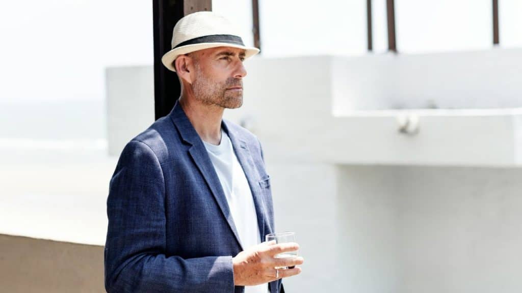 A man in a fedora, blue jacket, and white shirt stands outside holding a glass.