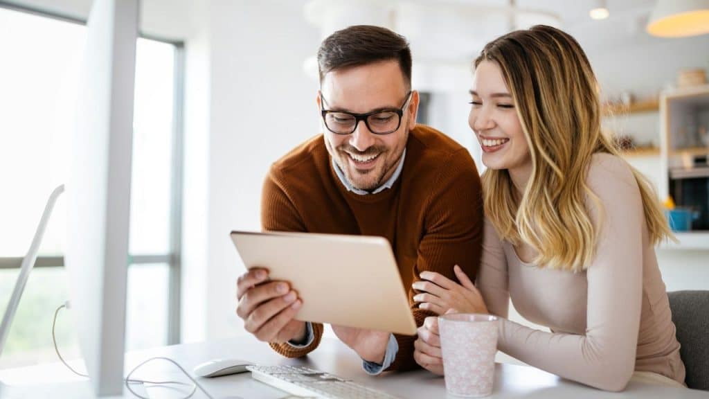 Smiling couple looking at a tablet together, with the woman holding a mug, in a bright modern office.