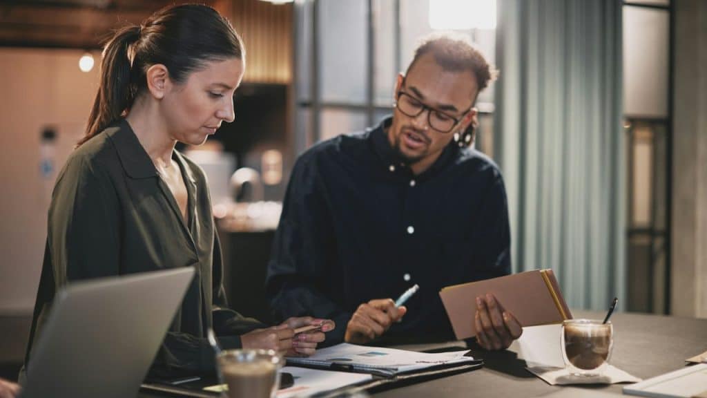 Two colleagues reviewing documents and a notebook at a table with coffee and a laptop in an office.