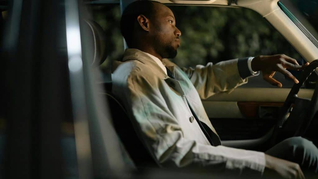A man in a light coat sits in a car at night, resting his arm on the steering wheel.