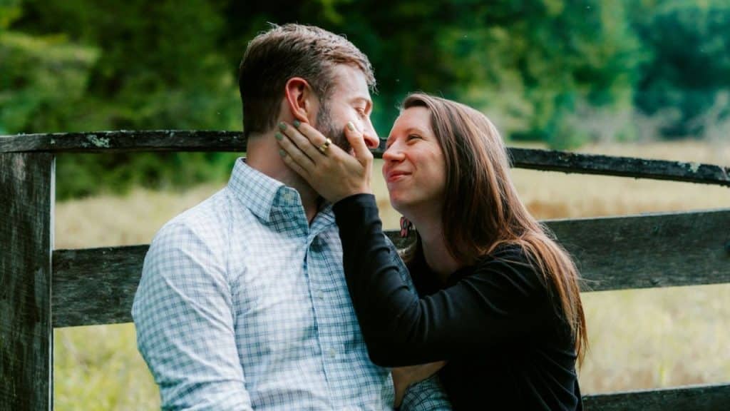 Woman touching a man's face and smiling at him outdoors near a wooden fence.