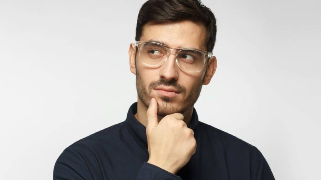 Man in clear glasses and dark shirt with hand on chin looking away thoughtfully.