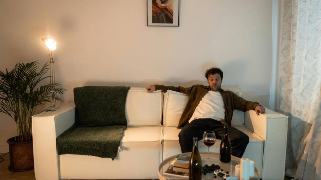 A man sitting on a couch with wine bottles and a glass on the table in front of him.