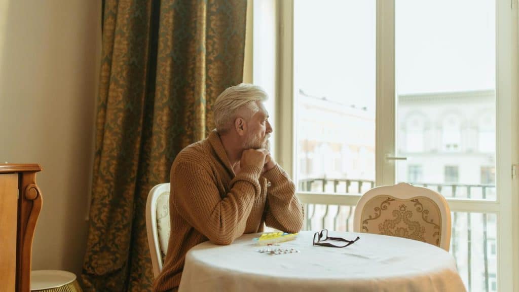 An elderly man sitting at a table and looking out the window.
