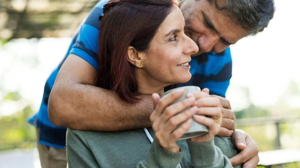 A man hugs a woman from behind as she holds a mug and smiles softly.