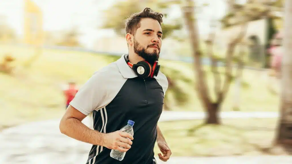 Bearded man with headphones around his neck running outdoors, holding a water bottle.