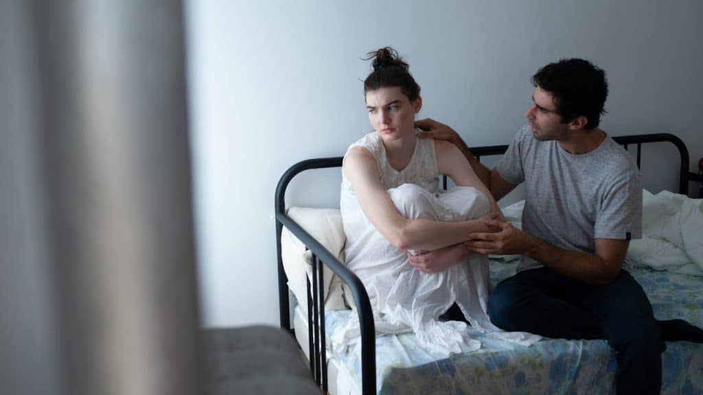 A man trying to console a woman who appears angry with him while seated on a bed.