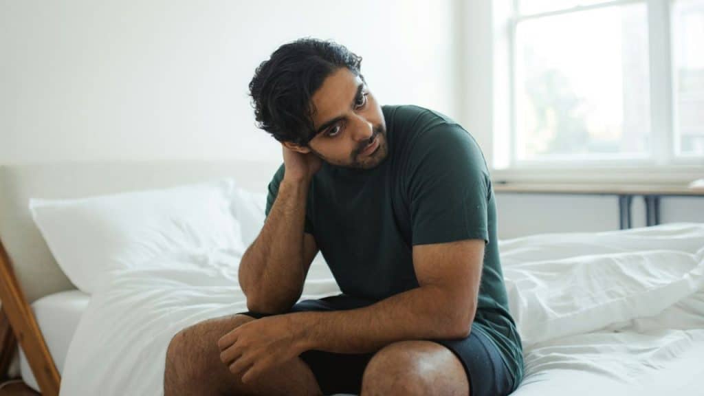 A concerned man in a dark green shirt sits on a white bed, resting his hand on his neck.
