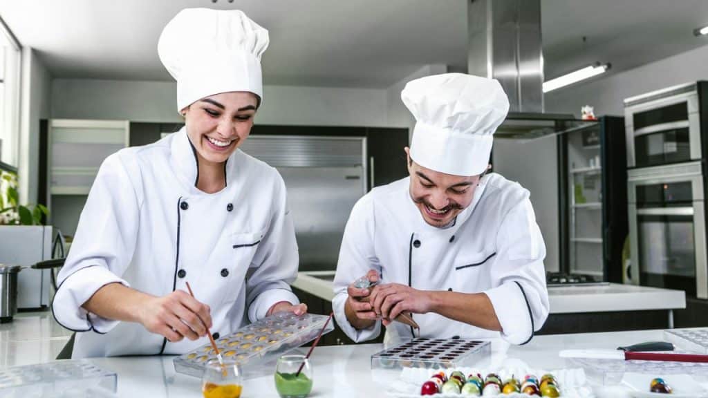 Two smiling chefs in white uniforms and toques are happily decorating chocolates in a bright, modern kitchen.