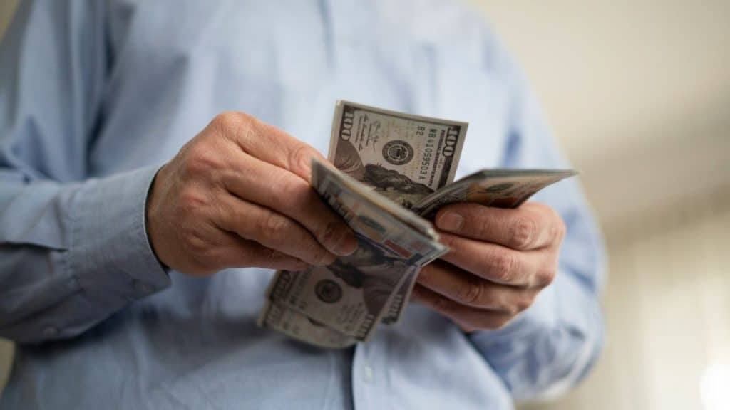 Close-up of a person in a light blue shirt counting a stack of US hundred-dollar bills.