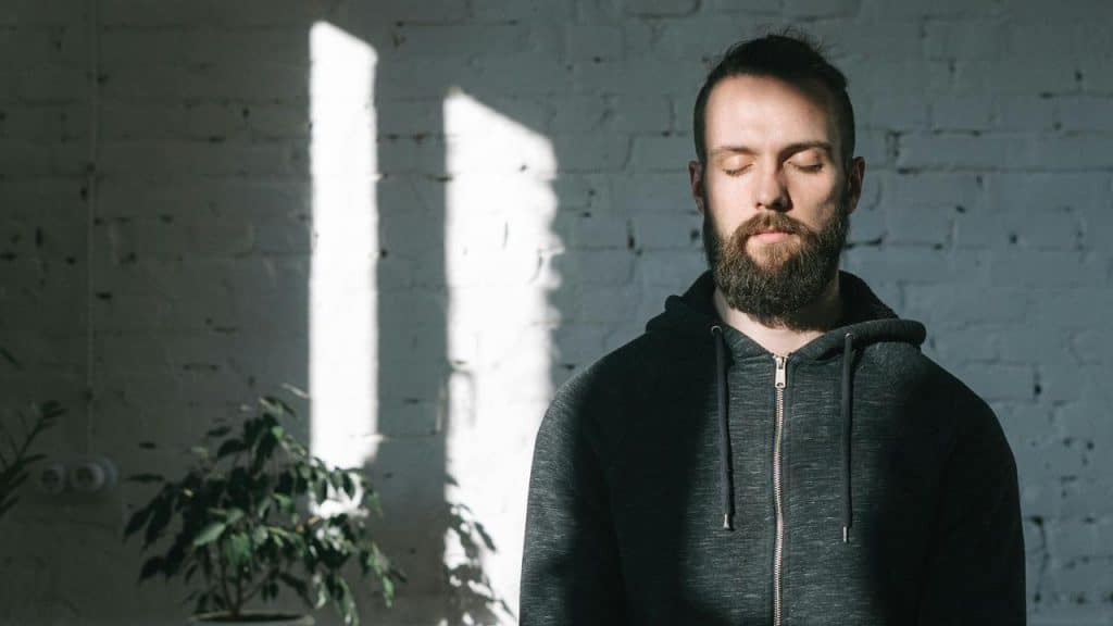 A man with closed eyes standing in sunlight against a brick wall.