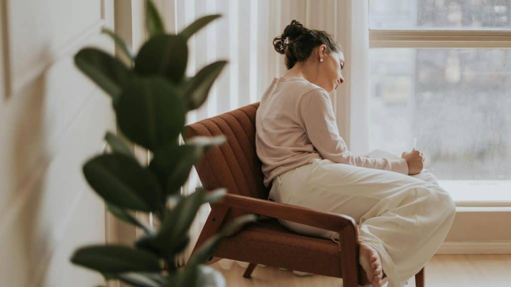 A woman sitting in a chair by the window writing in a notebook.
