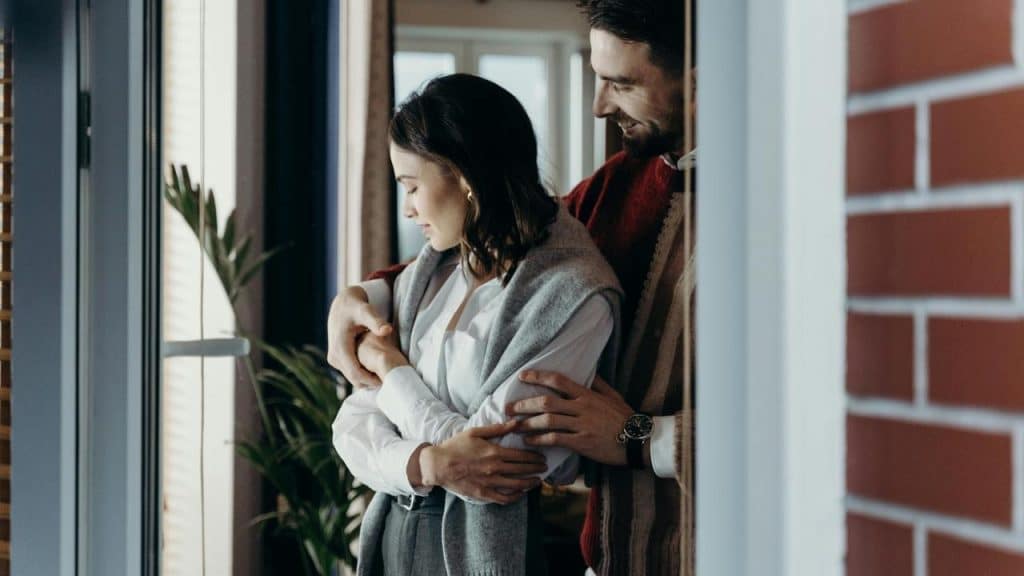 A man hugging a woman from behind near a window.