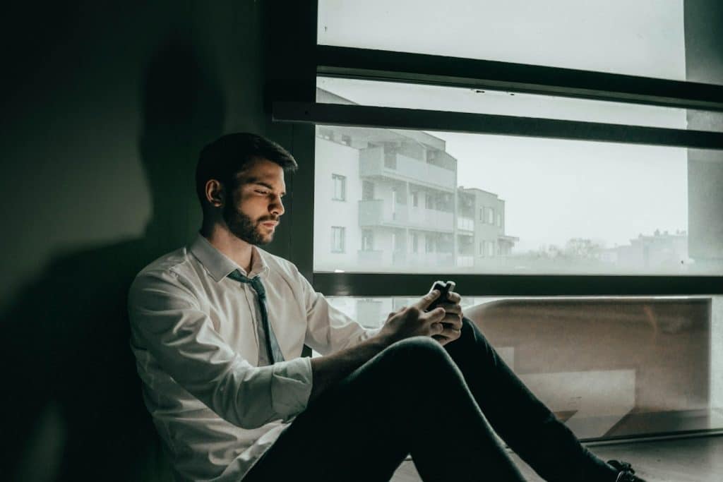 A man sitting beside the window, looking at his cellphone.