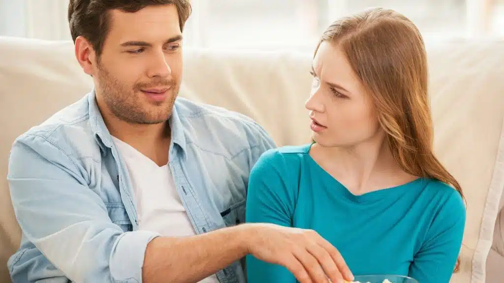 Young couple on a couch, man holding popcorn, woman looking at him with a frown or skeptical expression.
