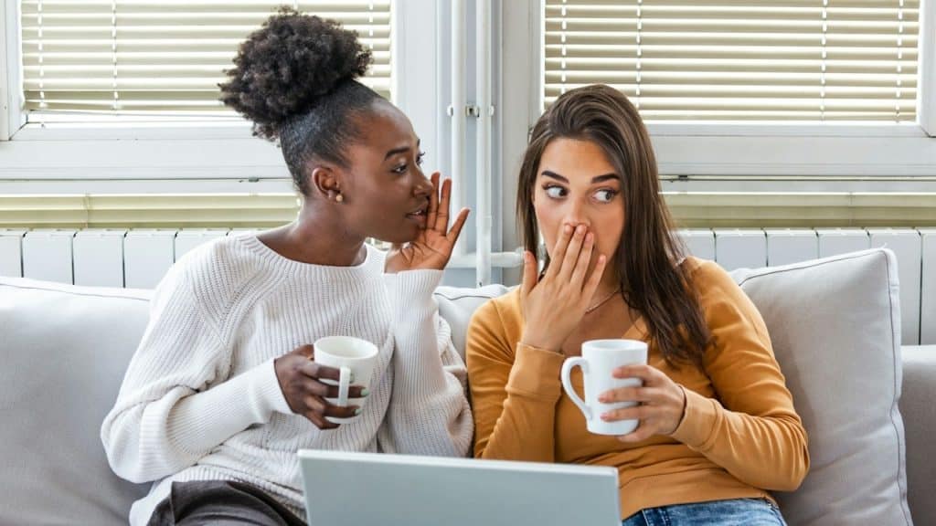 Black woman whispering a secret to a surprised woman with her hand covering her mouth, both sitting on a couch with mugs and a laptop.