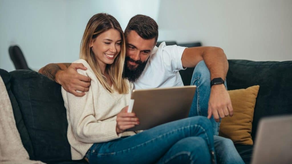 Smiling couple sitting on a couch, embracing and looking at a tablet together indoors.