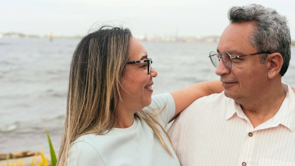 Smiling middle-aged couple in glasses outdoors, looking at each other with arm around shoulders.