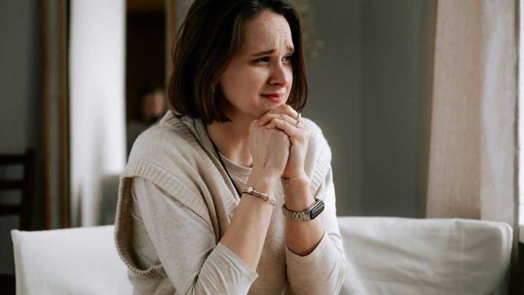 A woman with short hair sits, looking upset or distressed, with clasped hands.