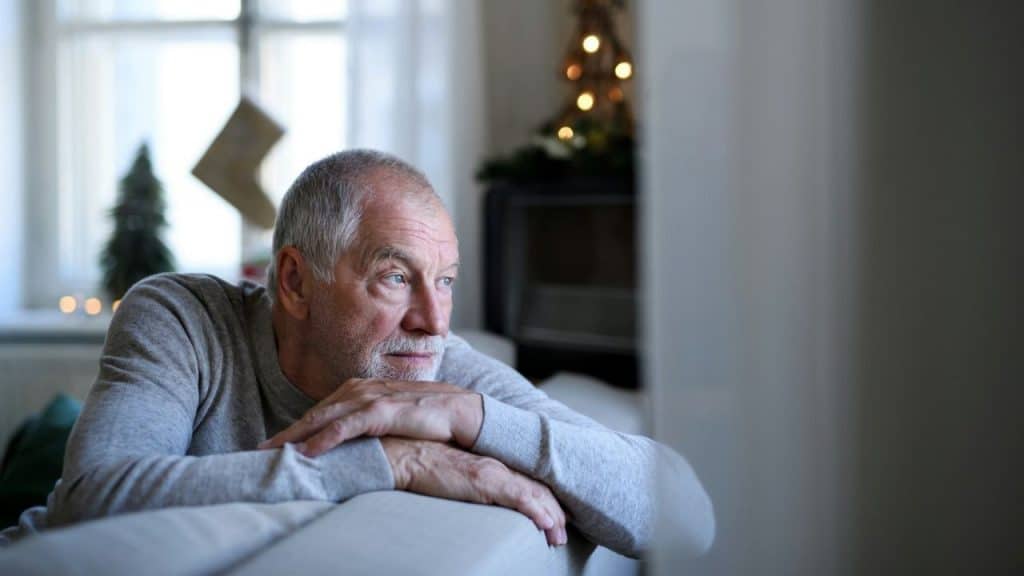 An elderly man in a gray sweater looks distant and pensive in a Christmas-decorated room.