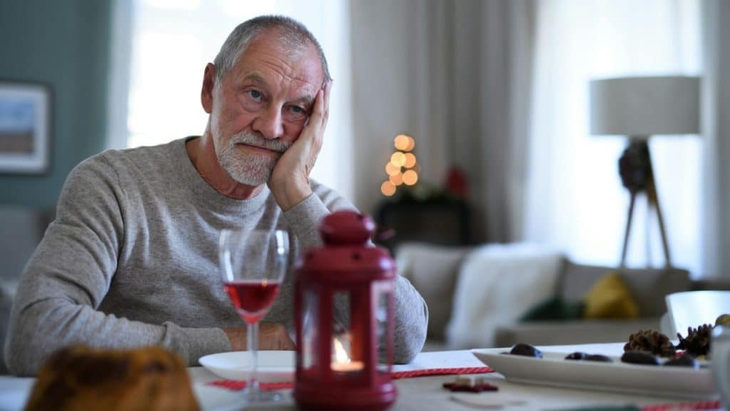 An elderly man sits alone at a table with a wine glass, looking sad or troubled.