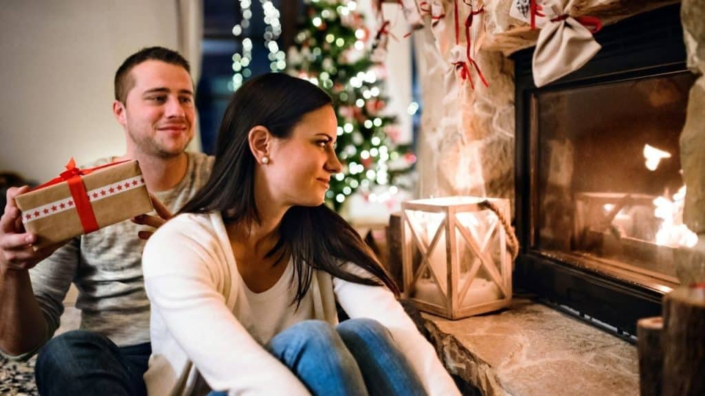 A couple sits by a fireplace at Christmas; the man holds a present.
