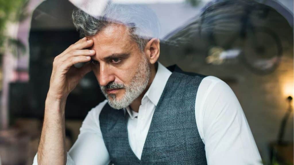 A middle-aged man in a vest and shirt stares through glass, resting his head on his hand.
