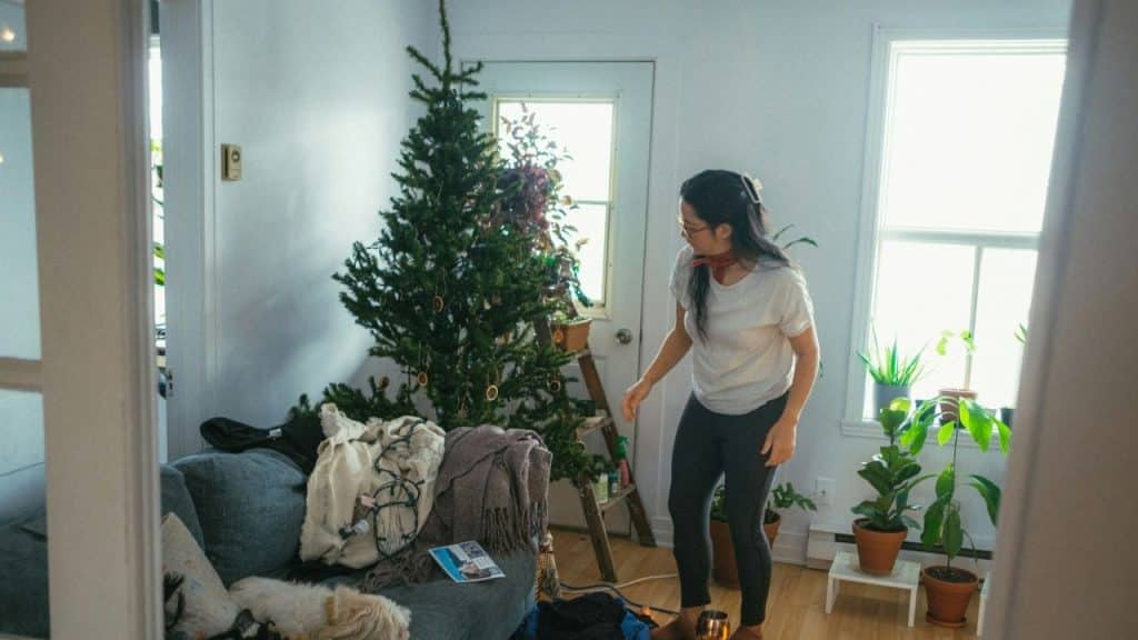 A woman stands in a bright room, looking distressed by a messy Christmas tree.