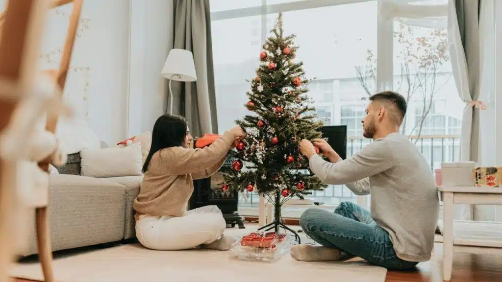 A man and woman sit on the floor, decorating a small Christmas tree together.