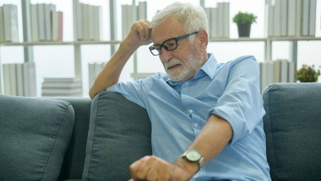 An elderly man in glasses and a light blue shirt sits distressed on a gray couch.