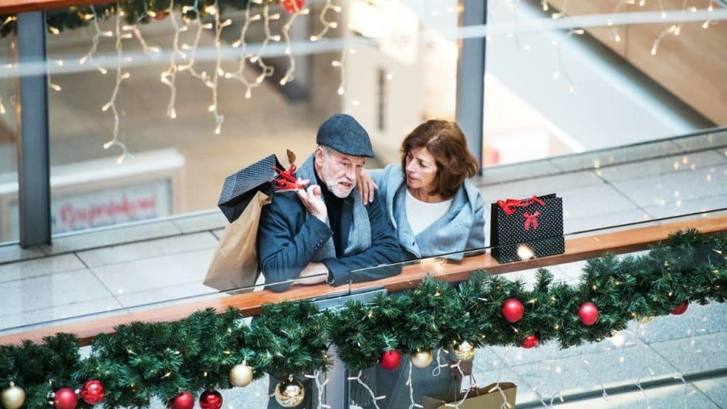 An older couple shops at a mall; the woman comforts the tired-looking man.